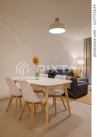 Dining area with white table, wooden legs, four white chairs and decorative vase in a cozy minimalist space Dining area with white table, wooden legs, four white chairs and decorative vase in a cozy minimalist space 125751834
