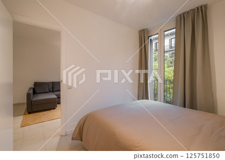 Bedroom interior with taupe bedspread, large window with beige curtains and view of living area Bedroom interior with taupe bedspread, large window with beige curtains and view of living area 125751850