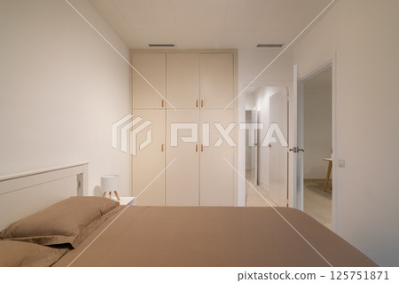Minimalist bedroom with taupe bedding, white built-in wardrobe and two open doorways Minimalist bedroom with taupe bedding, white built-in wardrobe and two open doorways 125751871