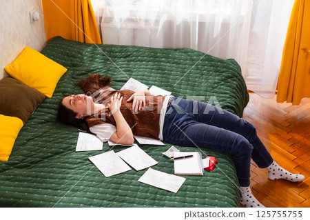 Young female organizing financial documents, calculating monthly costs, and planning household budget. Stressed woman lies on bed. Economy and finance. Mid shot 125755755