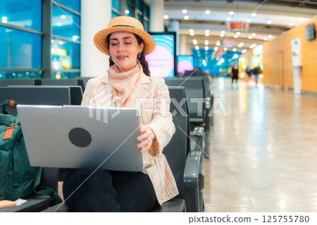 Mid shot of Caucasian elegant happy young woman is sitting on a chair in the waiting room of airport and working on a laptop. Copy space. Concept of remote work and travel 125755780