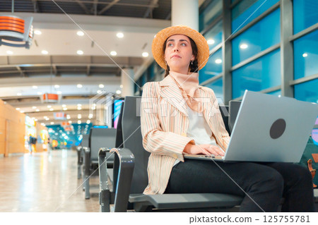 Bottom view of Caucasian elegant young woman is sitting on chair in waiting room of airport and working on laptop. Concept of remote work and travel 125755781
