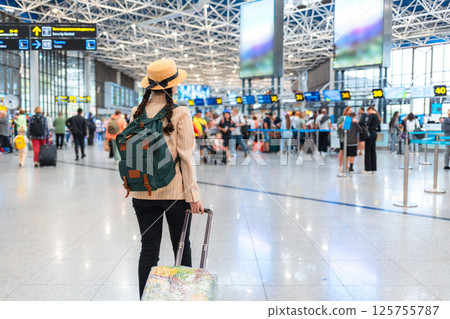 Rear view of elegant woman in a straw hat and backpack, wheeling a suitcase to the check-in desk. The concept of vacation and flight trip Rear view of elegant woman in a straw hat and backpack, wheeling a suitcase to the check-in desk. The concept of vacation and flight trip 125755787