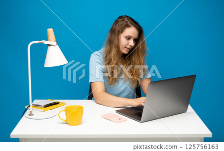 Woman working at a bright desk with laptop, notebook, and yellow mug on blue background 125756163