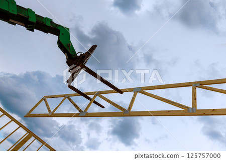 Telehandler machine at construction site lifts wooden truss into place with clouds scattered overhead. 125757000