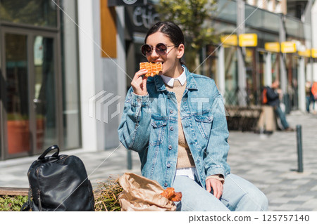 In a bustling city square, a 25-something woman enjoys a fresh waffle, capturing moments of solo travel, local cuisine, and relaxed lifestyle 125757140