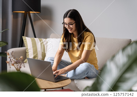 Young woman with glasses leans into her laptop, managing remote work deadlines while browsing for home items and checking in on her coursework 125757142