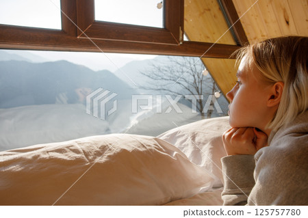 Pensive blonde woman laying on the bed and looking on the mountains in the window 125757780