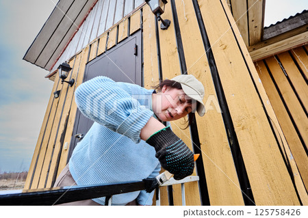 Woman uses paintbrush to coat metal porch railing with black paint aiming to restore and protect decorative elements of countryside home. Woman uses paintbrush to coat metal porch railing with black paint aiming to restore and protect decorative elements of countryside home. 125758426