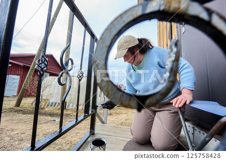 Woman performs detailed painting of metal porch railing, bending low to reach every spot as she applies black paint to refresh entrance of country home. 125758428