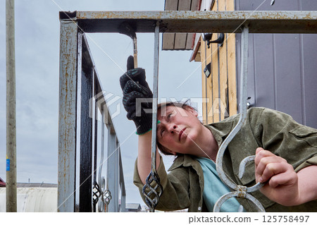 Painting process involves careful application of black paint with paintbrush on vertical scrolls of metal railing, woman appears focused and determined. Painting process involves careful application of black paint with paintbrush on vertical scrolls of metal railing, woman appears focused and determined. 125758497