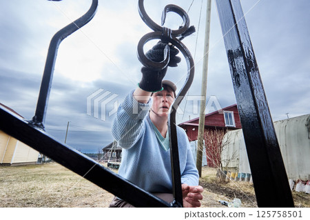 Gripping paintbrush with gloved hand, determined homeowner applies anticorrosive coating to porch rails, feeling satisfaction from visible progress. 125758501
