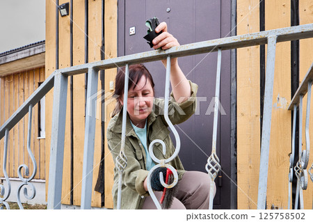 Removing rust from ornate porch railing, cheerful female painter uses wire brush with a smile, enjoying process of restoring metal to clean condition. 125758502