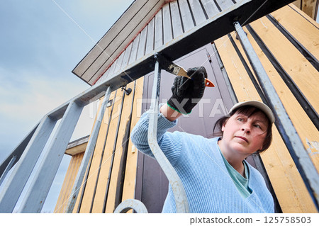 Applying protective paint to metal railings, woman carefully works on porch entrance of country house, focused on achieving smooth even coverage. Applying protective paint to metal railings, woman carefully works on porch entrance of country house, focused on achieving smooth even coverage. 125758503