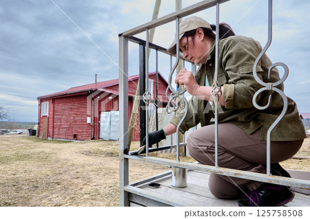 Middle aged Caucasian woman applies black paint to lower part of metal porch railing of private country house using paintbrush outdoors. Middle aged Caucasian woman applies black paint to lower part of metal porch railing of private country house using paintbrush outdoors. 125758508