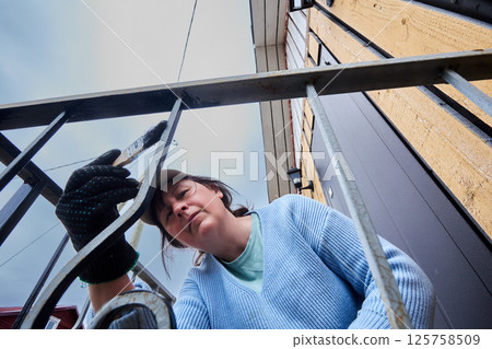 Careful strokes of paintbrush cover ornate metal bars of porch railing with black paint as woman concentrates on detailed painting near country home. 125758509