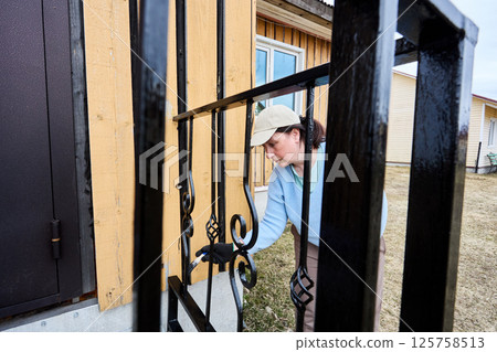 Leaning in close to metal railing, attentive house painter uses brush to cover every curve and angle, enjoying process of refreshing porch entrance. 125758513