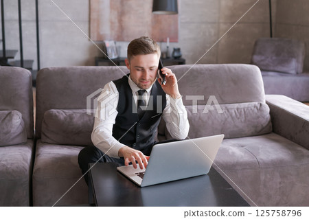A man in a formal suit is working on a laptop and conducting a telephone conversation in a cozy modern living room. 125758796