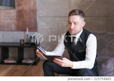 A young man in business clothes is writing something down in a notebook, sitting on a sofa in a modern interior. The concentrated look and formal style emphasize the professional setting 125758803