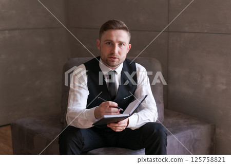 young man in business clothes is sitting on couch, intently making notes in notebook. The neutral background and strict style emphasize professional atmosphere and seriousness moment. young man in business clothes is sitting on couch, intently making notes in notebook. The neutral background and strict style emphasize professional atmosphere and seriousness moment. 125758821