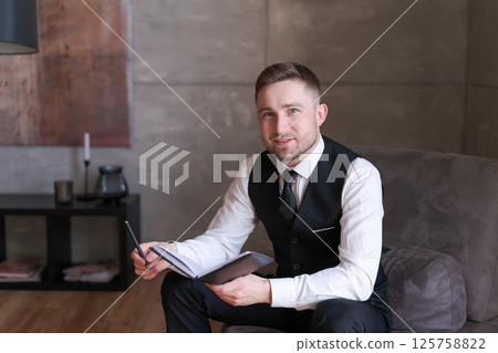 A man in a suit and vest holds a notebook and pen with a smile in an office setting, sitting on the couch. The lightness and confidence in his expression create an atmosphere of professionalism A man in a suit and vest holds a notebook and pen with a smile in an office setting, sitting on the couch. The lightness and confidence in his expression create an atmosphere of professionalism 125758822