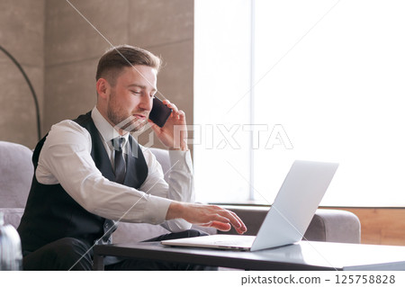 A man in a formal suit with a vest is sitting in a modern office, talking on the phone and working on a laptop, expressing confidence and professionalism A man in a formal suit with a vest is sitting in a modern office, talking on the phone and working on a laptop, expressing confidence and professionalism 125758828