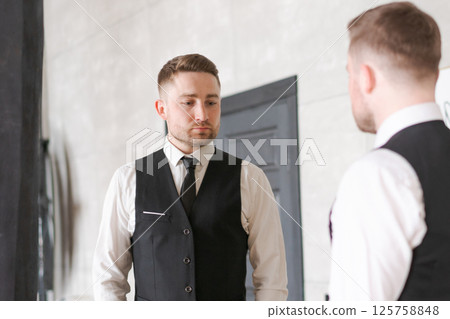 man in business clothes stands in front of a mirror, examining his image. The neutral background and soft lighting emphasize an atmosphere of concentration and introspection. man in business clothes stands in front of a mirror, examining his image. The neutral background and soft lighting emphasize an atmosphere of concentration and introspection. 125758848
