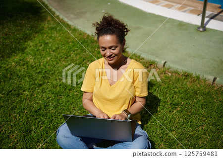 Smiling Woman Using Laptop Outdoors in a Green Park Setting 125759481