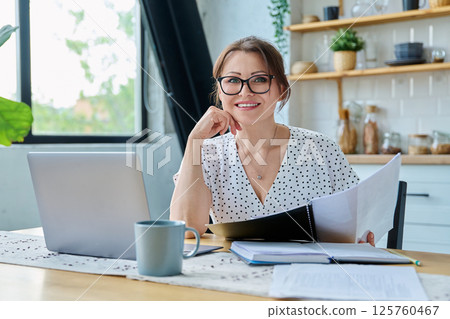 Middle aged smiling woman working with laptop, looking at camera, sitting at home Middle aged smiling woman working with laptop, looking at camera, sitting at home 125760467