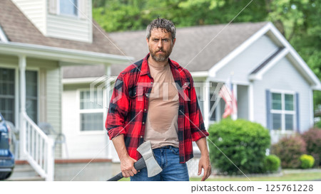 Lumberjack at backyard with an ax. Lumberjack with heavy axe. Strong sweating man after hard work. Lumberjack man with axe in front of the house. Lumberjack carries axe. Sweating man with axe 125761228