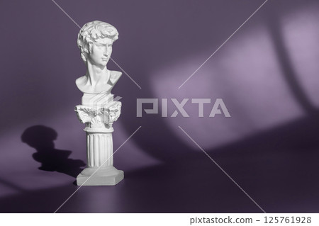 Gypsum copy of ancient statue head on purple background with light and shadow. White marble head of young man, head of famous statue by Michelangelo - David from Florence. Gypsum copy of ancient statue head on purple background with light and shadow. White marble head of young man, head of famous statue by Michelangelo - David from Florence. 125761928