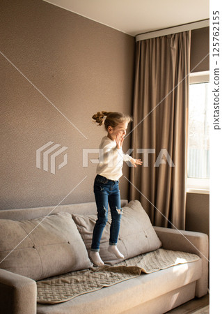 Blonde little girl get caught jumping on living room couch violating home rules. Candid shot of endless childhood energy. Image for parenting and childhood topics. Blonde little girl get caught jumping on living room couch violating home rules. Candid shot of endless childhood energy. Image for parenting and childhood topics. 125762155