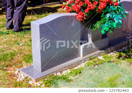 Attendees gather around gravestone adorned with flowers, honoring loved one in peaceful cemetery. 125762463