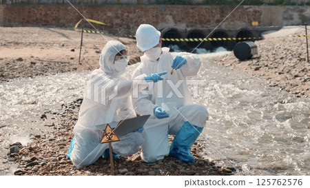 Biologist wear protective suit and mask collects sample of waste water from industry, problem environment, Ecologist sample taken dead fish to inspection and save data to laptop computer, Toxic water 125762576