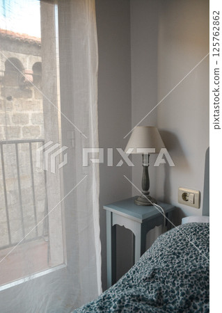 Bedside table with table lamp and beautiful vintage bed in hotel room with view of ancient fortress. Concept of adaptation of ancient architecture to modern needs Bedside table with table lamp and beautiful vintage bed in hotel room with view of ancient fortress. Concept of adaptation of ancient architecture to modern needs 125762862