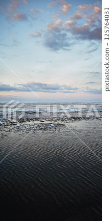 Spring landscape. Panoramic view of beautiful dawn on bay. Cumulus clouds over water in bright light. Ice, snow and rocks on coastline. Rising rays of sun are reflected in sea. Vertical photo. Spring landscape. Panoramic view of beautiful dawn on bay. Cumulus clouds over water in bright light. Ice, snow and rocks on coastline. Rising rays of sun are reflected in sea. Vertical photo. 125764031