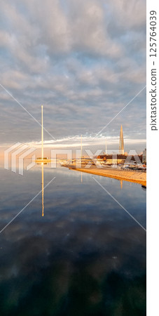 Spring landscape. Panoramic view of beautiful dawn on bay. Cumulus clouds over water in bright light. Ice, snow and rocks on coastline. Rising rays of sun are reflected in sea. Vertical photo. 125764039