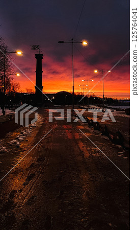 Fantastic winter landscape at fiery dawn. Silhouette of lighthouse and walking paths in park 300 anniversary of St. Petersburg, Russia. Vertical photo. Fantastic winter landscape at fiery dawn. Silhouette of lighthouse and walking paths in park 300 anniversary of St. Petersburg, Russia. Vertical photo. 125764041
