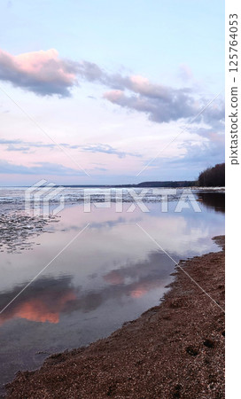 Spring landscape. Panoramic view of beautiful dawn on bay. Cumulus clouds over water in bright light. Ice, snow and rocks on coastline. Rising rays of sun are reflected in sea. Vertical photo. Spring landscape. Panoramic view of beautiful dawn on bay. Cumulus clouds over water in bright light. Ice, snow and rocks on coastline. Rising rays of sun are reflected in sea. Vertical photo. 125764053