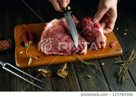 In a rustic kitchen, a chef expertly cuts fresh meat on a wooden cutting board. Spices and herbs are artfully arranged, highlighting the culinary process in action 125764366