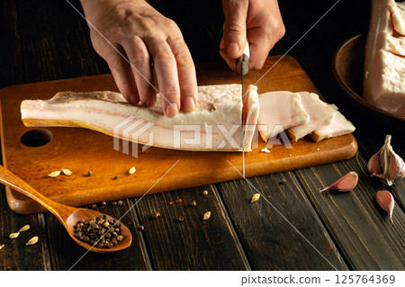 A chef skillfully slices pork belly on a wooden cutting board. Garlic and spices are arranged nearby, creating a rustic atmosphere in the kitchen. The focus is on the preparation of fresh ingredients 125764369
