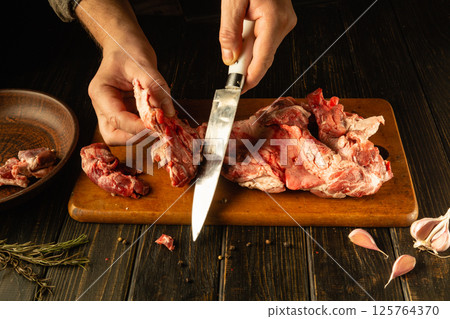 A person skillfully slices fresh meat on a wooden cutting board surrounded by garlic and herbs, creating a homely, culinary atmosphere in a rustic kitchen 125764370