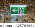 Illuminated Green and White Emergency Exit Sign with Arrow Pointing Left and Running Man Pictogram Mounted 125764951