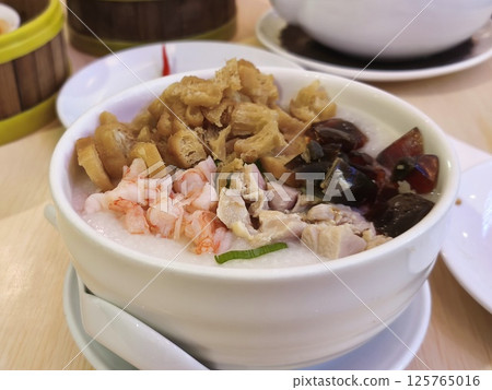 A bowl of congee topped with fried dough sticks, shrimp, chicken, and preserved black eggs, served in a white bowl with a spoon on a wooden table. 125765016