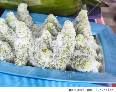 Kue lupis or an Indonesian steamed glutinosa rice cake snack, covered in shredded coconut and arranged on a blue tray with a banana leaf in the background. 125765136