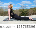 Women practicing yoga outdoors on sandy surface, performing upward facing dog pose on mat, enjoying sunny weather, clear sky, and natural surroundings for relaxation and wellness 125765396