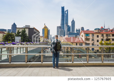A young man in a plaid shirt and jeans stands on a viaduct, gazing at Shanghai Pudong skyline with his backpack on, surrounded by blooming flowers 125767605