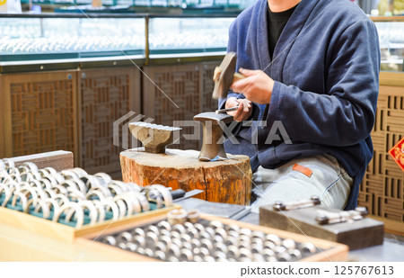 A craftsman in traditional blue jacket hand-forges jewelry rings on an anvil at Shanghai's Old Street Yunnan, with finished metal rings displayed in wooden trays at his open-air workshop 125767613
