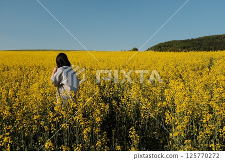 woman in white summer dress and straw hat walking through yellow rapeseed flower field under clear blue sky in countryside. High quality photo 125770272