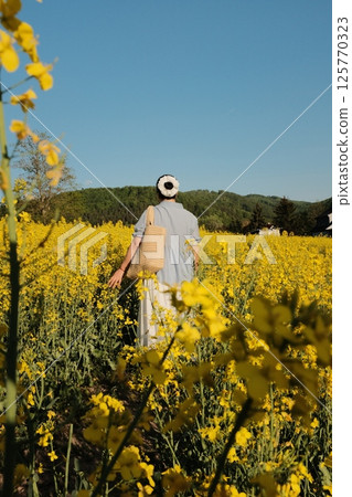 woman in white summer dress and straw hat walking through yellow rapeseed flower field under clear blue sky in countryside. High quality photo woman in white summer dress and straw hat walking through yellow rapeseed flower field under clear blue sky in countryside. High quality photo 125770323